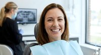 Happy middle-aged Caucasian woman patient smiling in dental chair during checkup visit at modern dentist office with female doctor in background.