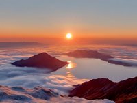 Majestic sunrise over mountain peaks piercing through sea of clouds, reflecting golden light on calm water surface and creating dramatic atmospheric landscape.