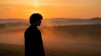 Young African American woman silhouette standing in misty landscape during golden hour sunrise. Contemplative moment with warm orange sky and rolling hills.