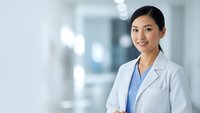 Young Asian female doctor in white lab coat smiling confidently in modern hospital corridor with arms crossed, representing healthcare professionalism.