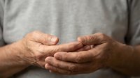Elderly man holding and massaging his hands, showing signs of joint pain or arthritis, close-up view against a casual gray background.