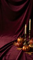 Elegant autumn still life with orange pumpkins, brass candlesticks with lit candles and oak leaves on luxurious burgundy velvet fabric background.