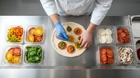 Professional chef in white uniform and blue gloves preparing fresh pizza with colorful toppings in commercial kitchen with ingredient containers.