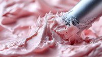 Pink buttercream frosting being spread with metal spatula on cake surface, showing smooth creamy texture for baking and dessert preparation.