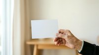 Senior male hand holding blank white card against neutral interior background with warm natural lighting, suitable for business presentations, marketing campaigns, and corporate messaging.