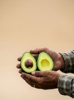 Hands holding two halves of fresh avocado with pit removed against neutral background. Suitable for healthy eating, nutrition, organic produce, farming, and food marketing campaigns.