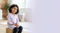 Young girl with curly dark hair and medium skin tone sitting on woven pouf, wearing lavender patterned shirt and dark pants, looking at camera in bright modern interior space.