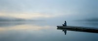 Solitary figure sitting on wooden dock over calm misty lake at dawn, peaceful morning reflection scene for meditation and tranquility concepts.