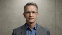 Professional middle-aged Caucasian businessman in gray suit and blue shirt wearing glasses against concrete wall background for corporate headshot.