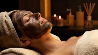 Young Caucasian man relaxing with chocolate facial mask treatment at luxury spa salon with candles and aromatherapy diffuser in warm ambient lighting.