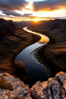 Dramatic sunset over winding river canyon with golden light illuminating rocky cliffs and dramatic clouds, reflecting warm colors on meandering water surface below.