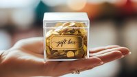 Small transparent gift box with golden treats held in open palm against blurred festive background for holiday gifting and special occasions.