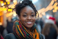Young African American woman with natural hairstyle wearing colorful knitted scarf smiling outdoors against blurred holiday lights and festive background.