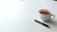 Steaming cup of tea with black pen on clean white desk surface creating minimalist workspace atmosphere for productivity and relaxation moments.