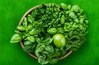 Fresh green herbs and apple arranged in wooden bowl on bright green background, overhead view of aromatic culinary ingredients for cooking and garnishing.