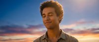 Young mixed-race man with curly hair smiling peacefully with closed eyes during golden hour sunset. Serene expression against colorful sky for wellness concepts.