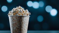Fresh popcorn overflowing in white paper container against dark background with soft blue bokeh lights, creating cinematic atmosphere for movie night concept.