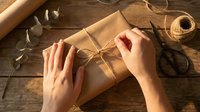 Hands wrapping kraft paper gift box with twine on wooden table, surrounded by scissors, ribbon and craft supplies for present packaging and decoration.