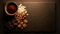 Cocoa powder in wooden bowl with almonds, walnuts, chocolate pieces and coconut flakes arranged on dark slate board for baking and cooking recipes.