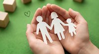 Hands protecting white paper cutout family figures on green background with wooden blocks. Family insurance, care and protection concept for healthcare design.