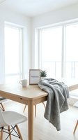 Minimalist Scandinavian dining room with wooden table, gray throw blanket, welcome sign, and plant in bright white interior.