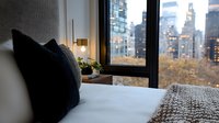 Cozy bedroom interior with city skyline view through window at dusk, featuring white bedding, knit throw blanket, decorative pillows and warm table lamp creating intimate atmosphere.