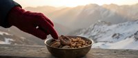 Person in red knit glove holding bowl of nuts with snow-covered mountains at sunrise, outdoor winter lifestyle scene.