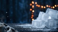 Stacked ice cubes with sparkling frost on dark surface, warm bokeh lights and snowflakes in blurred background, suitable for beverage, winter and cooling concepts.