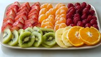 Fresh sliced fruits arranged in colorful rows on white plate including strawberries, mandarin oranges, raspberries and kiwi creating vibrant healthy food composition.