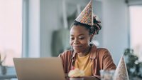 Young African American woman wearing party hat smiling at laptop during virtual celebration in bright modern office space.