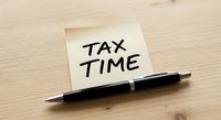 Yellow sticky note with tax time reminder message and black pen on wooden desk surface for financial planning and accounting preparation.