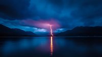 Lightning bolt strikes between mountains, reflecting on calm lake surface under dramatic night sky with purple and blue storm clouds. Perfect for weather themes.