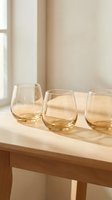 Three elegant amber tinted stemless wine glasses arranged on natural wood table in soft sunlight creating warm shadows for home decor and entertaining.