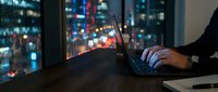 Professional working late on laptop computer with city lights bokeh background through office window at night.