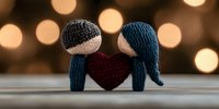 Handmade knitted couple dolls with burgundy heart between them against blurred warm bokeh lights background, romantic Valentine's day concept.