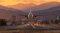 Commercial airplane landing at airport runway during golden hour sunset with mountain landscape and city lights creating dramatic aviation scene for travel industry.