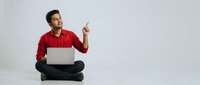 Young Indian man in red shirt sitting cross-legged with laptop pointing up, excited expression on white background for business and technology concepts.