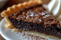 Rich chocolate fudge pie slice with flaky golden crust and dusting of cocoa powder on white plate, extreme close up showing moist, dense texture and glistening surface.