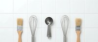 Kitchen utensils arranged in row on white tile background including whisks, measuring spoons and pastry brushes for cooking and baking preparation.