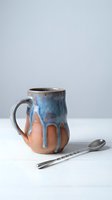Handmade ceramic mug with blue dripping glaze over terracotta clay body, artisan pottery coffee cup with metal spoon on white surface for kitchen decor.