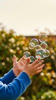 Child's hands in blue sleeve catching iridescent soap bubbles outdoors with blurred green foliage and golden bokeh background, capturing playful summer moments.