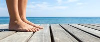 Close-up of bare feet on wooden boardwalk overlooking calm ocean and blue sky, summer vacation and relaxation concept for travel and leisure design projects.