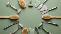 Spa wellness tools arranged in circle on green background including jade rollers gua sha stones wooden brushes and metal beauty instruments for skincare routine.