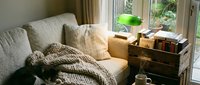 Cozy reading nook with neutral upholstered sofa, layered knit blankets, wooden side table with books and green lamp, natural window light, warm home interior design.