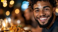 Young African American man with bright smile and beard enjoying gourmet meal at upscale restaurant with festive bokeh lights in background, expressing joy and satisfaction.