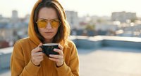 Young Caucasian woman in yellow hoodie and sunglasses holding smartphone on urban rooftop during golden hour sunset with city skyline background.