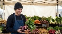 Young male farmer in beanie and apron holding fresh beets at outdoor farmers market with organic vegetables displayed under tent for healthy local food business.