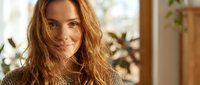 Young Caucasian woman with long wavy blonde hair smiling warmly in cozy knit sweater at home with natural sunlight and plants in background for lifestyle content.