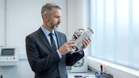 Middle-aged Caucasian male professional businessman in dark suit and tie holding industrial robotic hand in modern office laboratory setting with windows and equipment.
