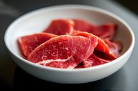 Fresh raw beef slices arranged in white ceramic plate on dark background, showing marbled texture and rich red color. For food photography and culinary use.